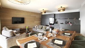 A modern living room with a wooden floor, grey sofas, a wooden dining table set for six, and a kitchen area with dark cabinets and a flat-screen TV mounted on the wall. - Magnifique vue sur les pistes, exposé sud, garage (Alpe d'Huez)