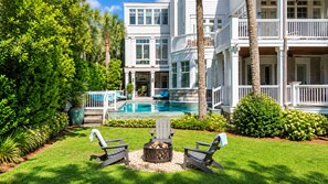 A pool area with lounge chairs, a fire pit, and a large building with balconies. - Luxury Beachfront Home with Guesthouse, Heated Pool & Spa, Putting Green, East Beach Oasis! (Saint Simons Island)