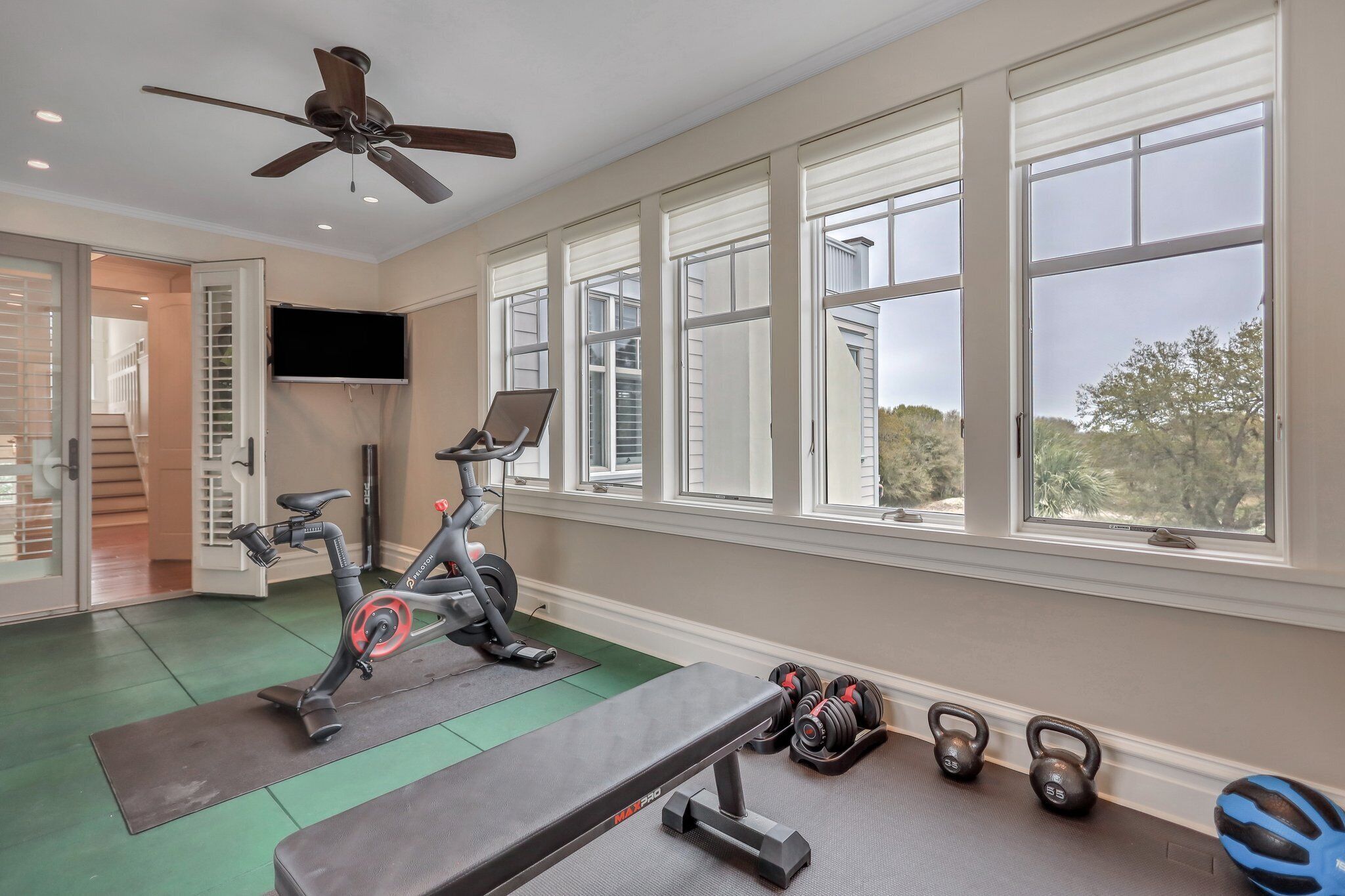 Ein Heimfitnessraum mit Trainingsgeräten, großen Fenstern und einem Deckenventilator.
