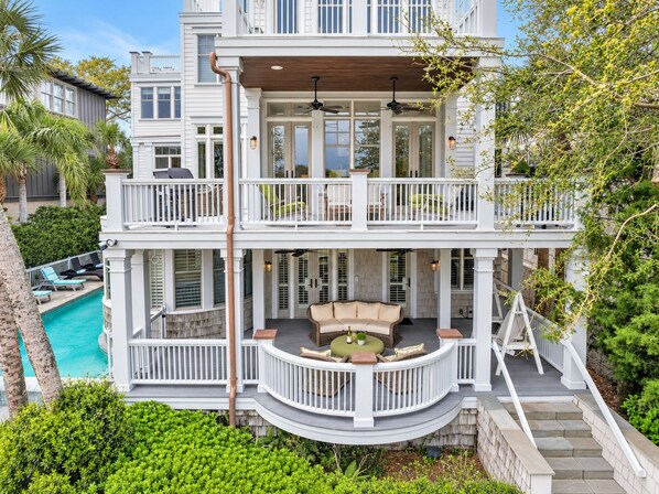 A two-story house with a balcony, a pool, and a patio with outdoor furniture. - Luxury Beachfront Home with Guesthouse, Heated Pool & Spa, Putting Green, East Beach Oasis! (Saint Simons Island)