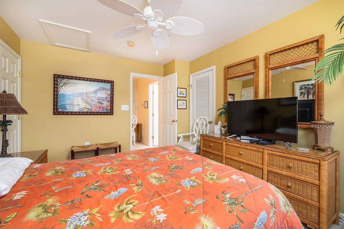 A hotel room with a bed, a television, a dresser, a fan, and a painting on the wall.
