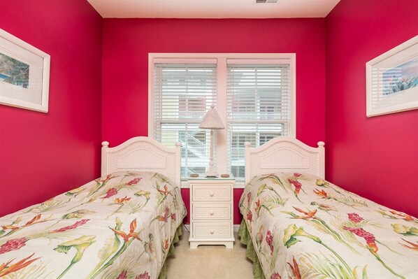 A room with two beds, a window with blinds, a bedside table, and a lamp.