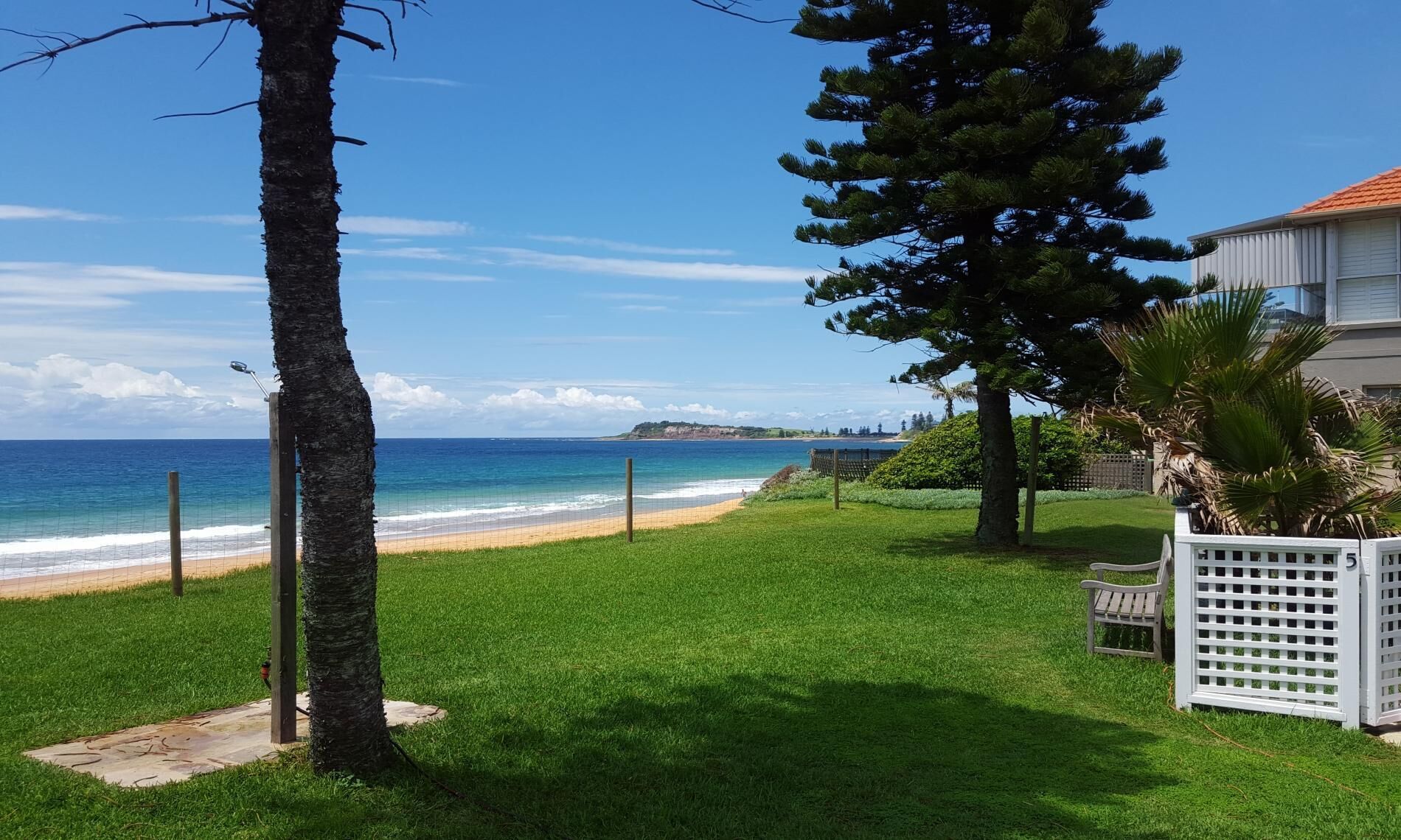 Une zone herbeuse avec une plage et une vue sur l’océan, une clôture blanche et une maison avec un toit rouge.