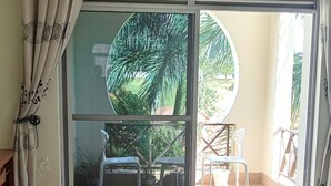 Une chambre à coucher avec un lit, une grande fenêtre avec des rideaux, une vue sur un balcon avec des chaises et un sol carrelé.