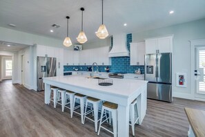 A modern kitchen with white cabinetry, a large island with bar stools, stainless steel appliances, and blue-tiled backsplash. - Endless Summer (Bradenton Beach)