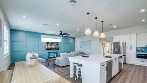Una cocina moderna con armarios blancos, una isla central con taburetes, un ventilador de techo, lámparas colgantes y vistas al área de estar con una pared azul y un sofá.