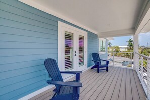 A covered porch with two Adirondack chairs, a white door with shutters, and a view of a parking lot and palm trees. - Endless Summer (Bradenton Beach)