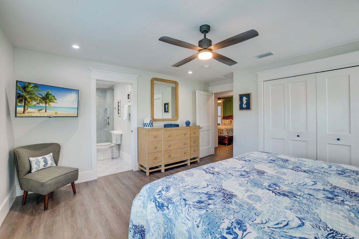 A bedroom with a bed, a chair, a dresser, a ceiling fan, and a painting on the wall.