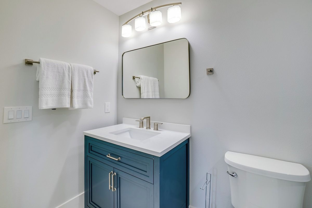 A bathroom with a white sink, a blue cabinet, a white toilet, and a wall-mounted mirror with lights.