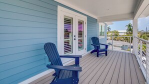 Un porche cubierto con dos sillas Adirondack, una puerta blanca con contraventanas y vistas al océano y palmeras.