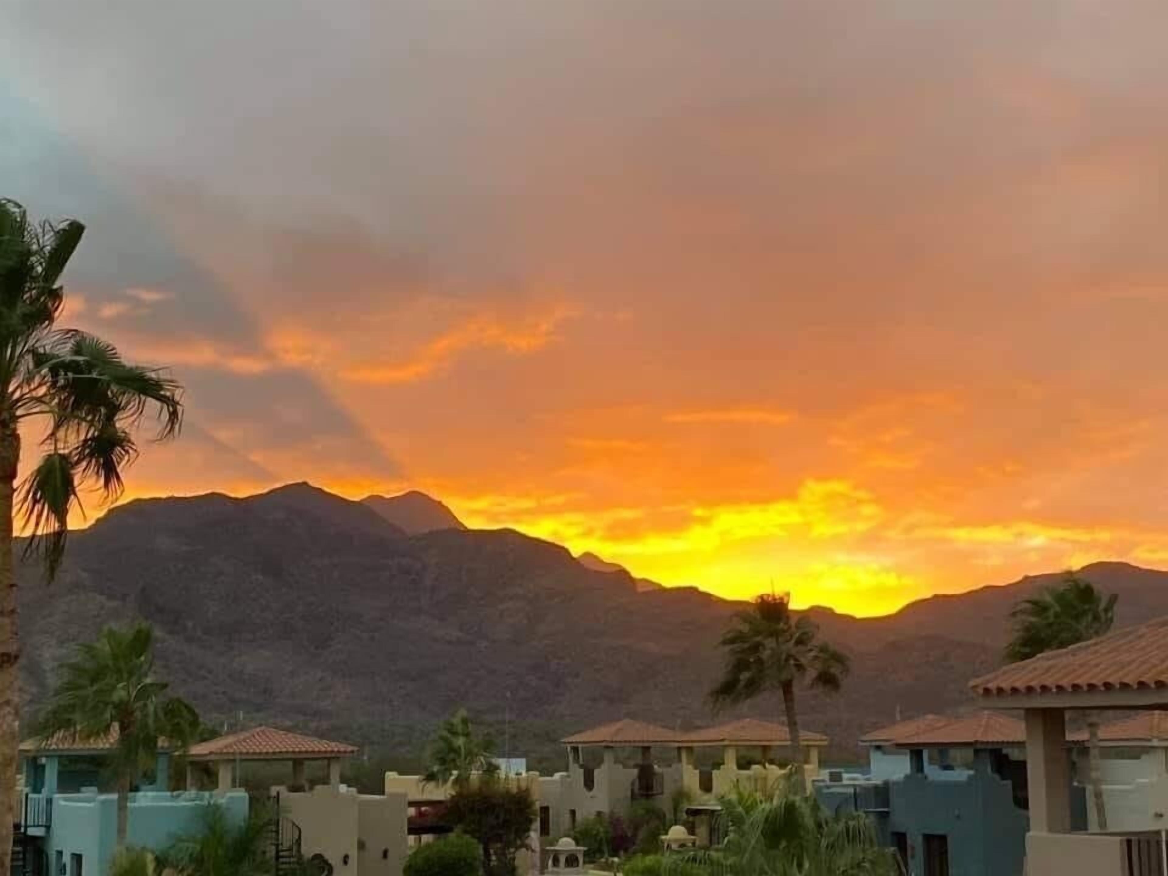 A residential area with houses and palm trees, set against a backdrop of mountains during sunset.