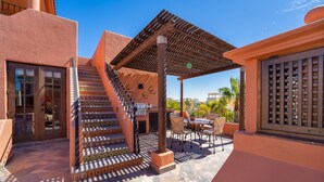 Un espacio al aire libre cubierto con una pérgola de madera, una escalera con baldosas estampadas y una zona de estar con sillas y una mesa.