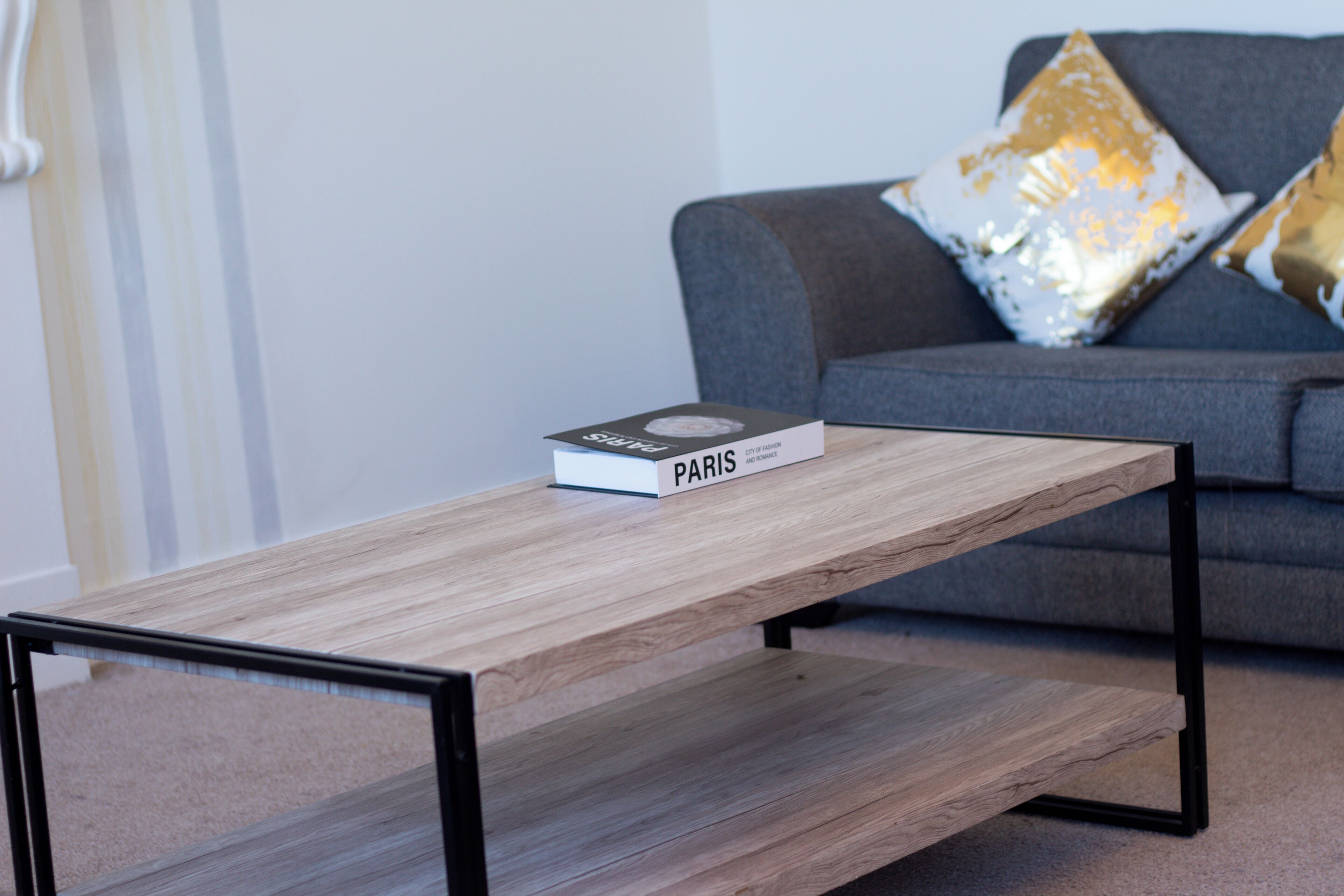 A modern living room with a wooden coffee table, a book titled "PARIS," and a grey sofa with decorative pillows.