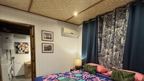 Un dormitorio con cama, mesita de noche, lámpara, aire acondicionado instalado en la pared y puerta que da a un baño.