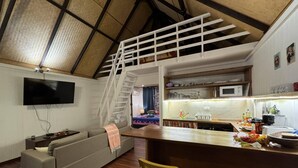 Una sala de estar moderna con un sofá gris, una mesa de comedor de madera con dos sillas, una zona de cocina con microondas, y una escalera que conduce a un nivel superior.