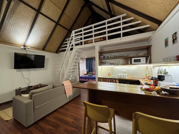 A modern living room with a grey sofa, a wooden dining table with two chairs, a kitchen area with a microwave, and a staircase leading to an upper level. - Iinui Legends Travels (Moorea-Maiao)