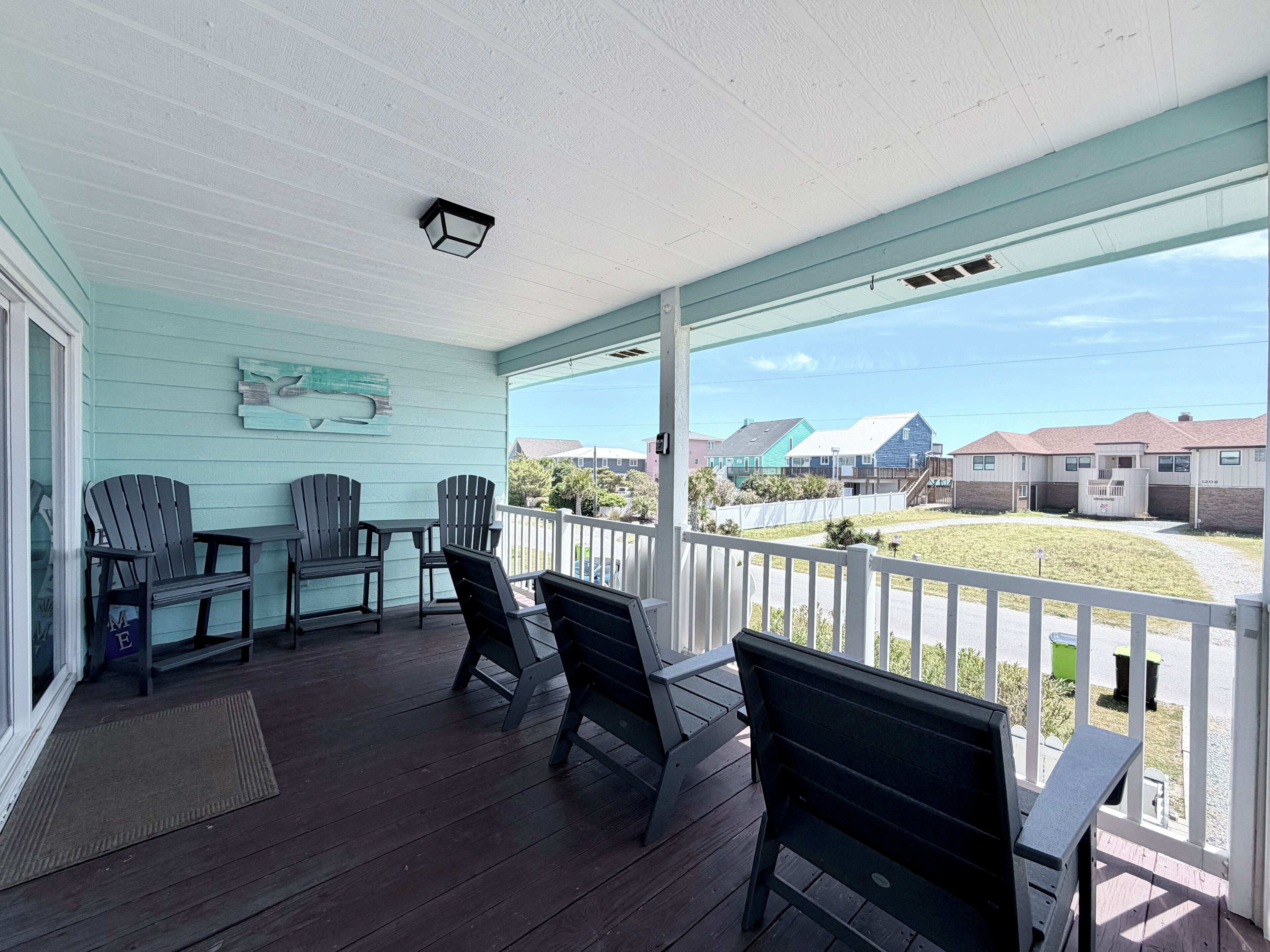 A covered porch with wooden flooring, several Adirondack chairs, and a view of a residential area with houses and a clear sky.