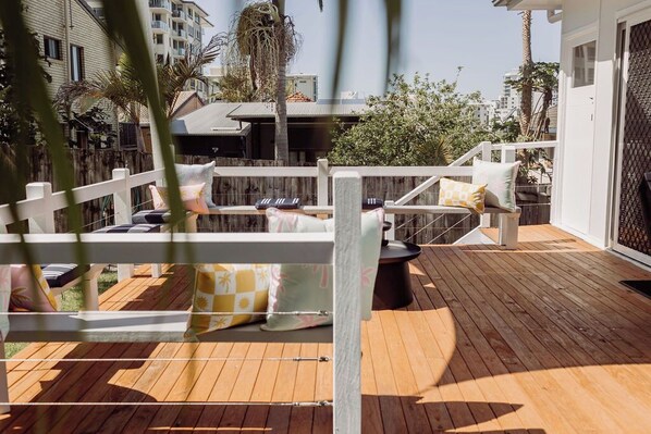 Une terrasse en bois avec une clôture blanche, des sièges d’extérieur agrémentés de coussins et une vue sur les bâtiments en arrière-plan.