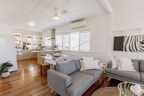 Un salon moderne avec un canapé gris, une table à manger et un coin cuisine doté de meubles blancs et d’un ventilateur de plafond.