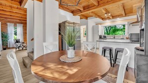 A modern kitchen with wooden beams, a central island, and a dining area with a round table and white chairs. - Wooded 3BR Luxury Cabin, Close to City & Nature (Greenville)