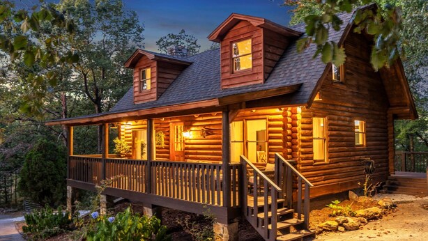 A log cabin with a porch, a staircase, and a well-lit interior. - Wooded 3BR Luxury Cabin, Close to City & Nature (Greenville)