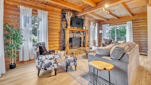 A cozy log cabin interior with a stone fireplace, wooden ceiling beams, and a comfortable seating area. - Wooded 3BR Luxury Cabin, Close to City & Nature (Greenville)