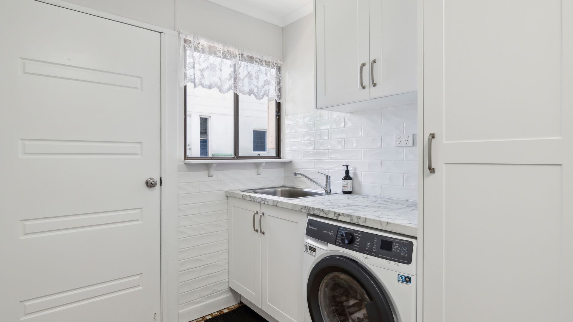 A compact laundry room with a washing machine, a sink, and a window with a lace curtain.