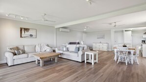 Amplia sala de estar con un sofá blanco, una mesa de centro y una mesa de comedor con sillas. Se observa un área de cocina con gabinetes blancos y una mesa de comedor.