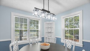 A dining room with a dark wooden table, white chairs, and a bowl of bread. There are two windows with blinds, a white decorative shelf, and a hanging light fixture. - Forest Hills Manor w/ King Bed (Augusta)