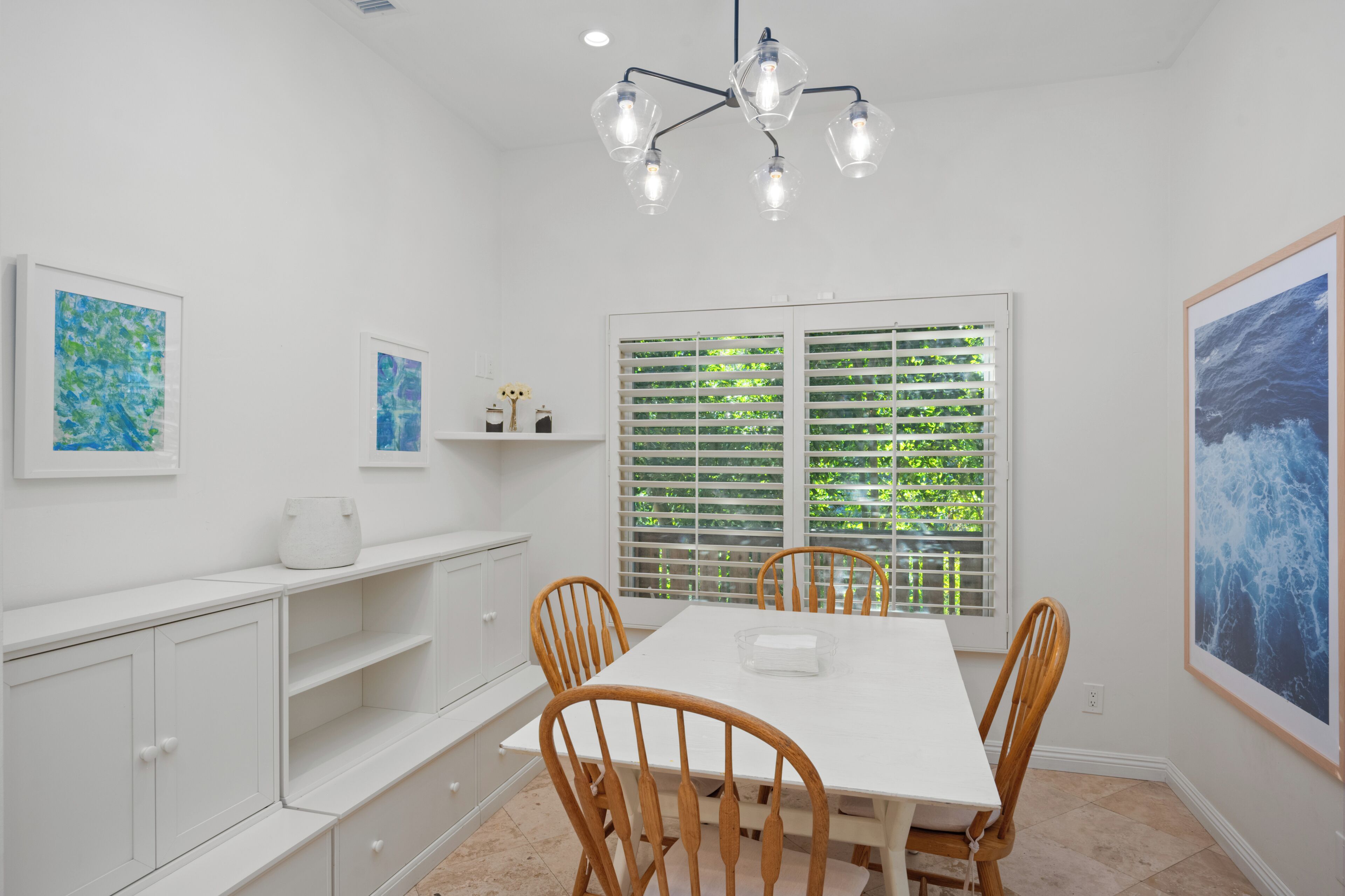 Un coin repas avec une table blanche, des chaises en bois, un lustre et des œuvres d’art accrochées aux murs.