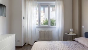 Ein Schlafzimmer mit Bett, Heizkörper, Fenster mit Vorhängen, Nachttisch mit Lampe und einem an der Wand befestigten Fernseher.