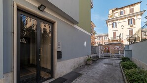 Ein Gebäudeeingang mit Glastür, ein kleiner Außenbereich mit Metallgitter und eine Treppe, die zu einem Balkon führt.