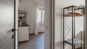 Ein Zimmer mit Holzboden, einer Küchenzeile mit Herd, einem Heizkörper und einem Fenster mit Vorhängen. An der Wand befindet sich ein Metallregal mit hölzernen Kleiderbügeln.