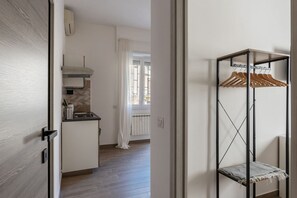 Una habitación con piso de madera, una zona de cocina con estufa, un radiador y una ventana con cortinas. En la pared, hay un estante de metal con perchas de madera.