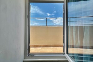 Een kamer met een raam dat uitkijkt op een balkon met uitzicht op de lucht en wolken.