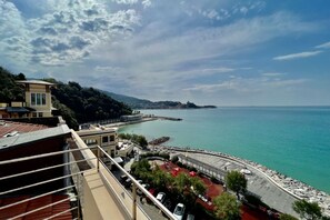 A coastal view with a building overlooking the sea, a parking area, and a promenade with red flooring.