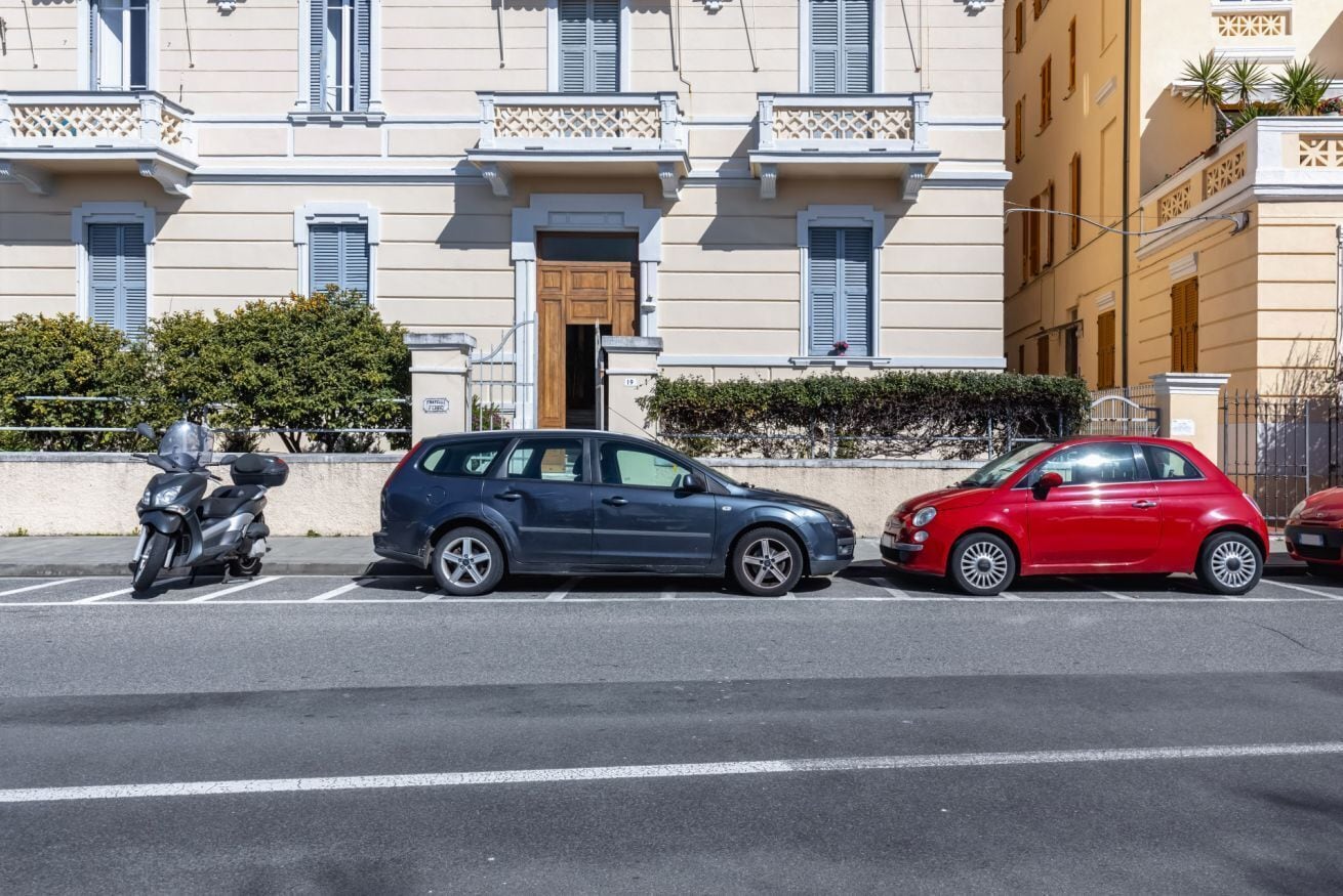 A street with parked cars and a motorcycle, buildings with balconies and shutters, and a clear sky.