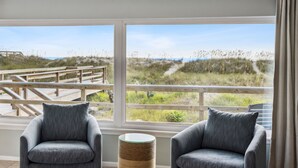 Una habitación con dos sillones grises, una mesa redonda de mimbre y un amplio ventanal con vista a una playa y una terraza de madera.