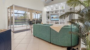 Un salon spacieux avec un canapé vert, un ventilateur de plafond et une grande fenêtre donnant sur une piscine.