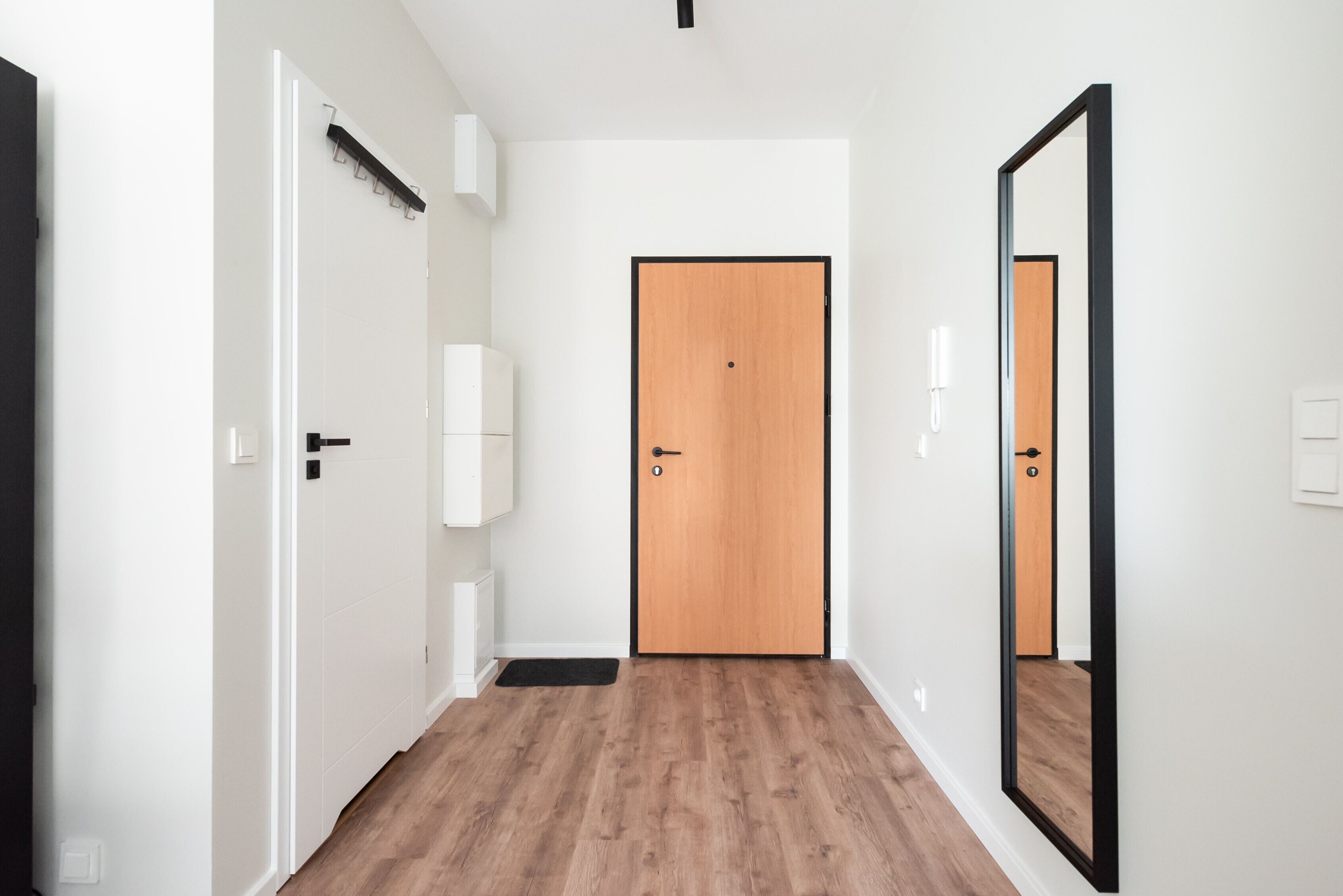 A hallway with a wooden floor, white walls, a wooden door, and a black-framed mirror.