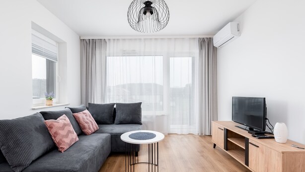 Een moderne woonkamer met een grijze bank, een ronde salontafel, een televisie op een houten dressoir, en een groot raam met transparante gordijnen.