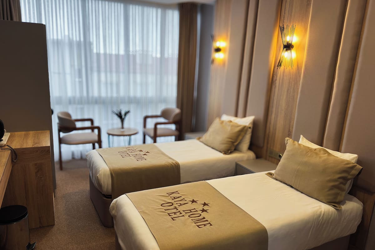 A hotel room with two beds, a small table, chairs, a lamp, and a view of the outside through sheer curtains.