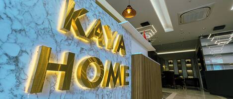 Un'elegante hall d'hotel moderna, con una parete in marmo e un'ampia insegna illuminata con la scritta "KAYA HOME".