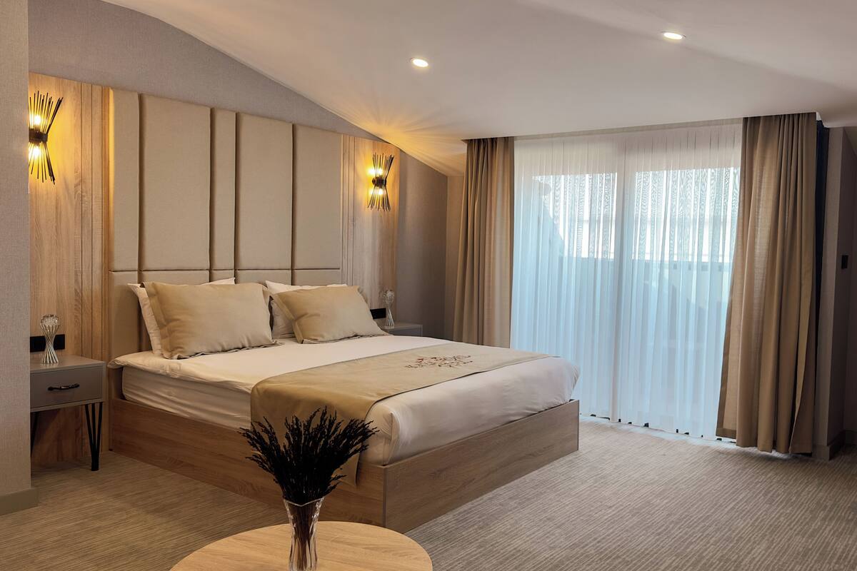 A modern hotel room with a large bed, a bedside table, a nightstand, a desk, and a large window with curtains.