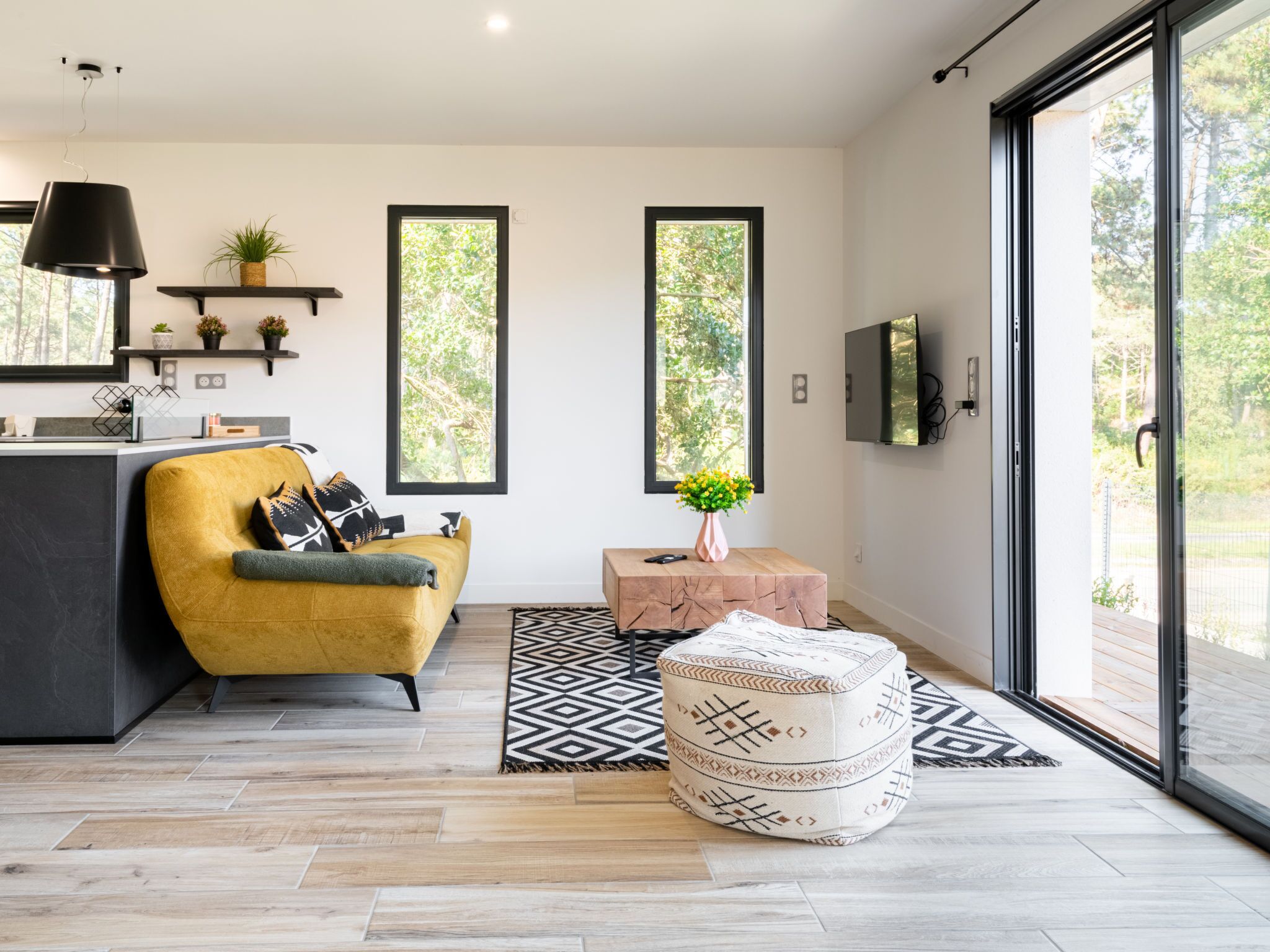 A modern living room with a yellow sofa, a coffee table, and a patterned ottoman. There are two windows with views of trees, a flat-screen TV on the wall, and a pendant light above the sofa.