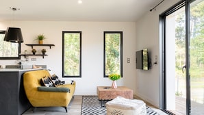 Ein modernes Wohnzimmer mit einem gelben Sofa, einem Couchtisch und einem gemusterten Hocker. Zwei Fenster bieten Blick auf Bäume, ein Flachbildfernseher ist an der Wand angebracht und über dem Sofa hängt eine Pendelleuchte.