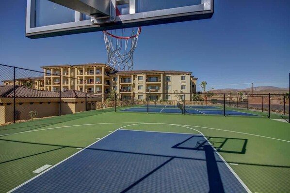 An outdoor basketball court with a net, surrounded by a fence, and buildings in the background.