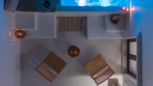 A room with a large window, a bed, a small table, a chair, a plant, and a heater. - Electra Suites & Spa (Santorini)
