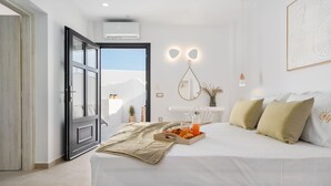 A modern bedroom with a large bed, a bedside table with a tray of oranges and juice, and a view of the mountains through an open door. - Electra Suites & Spa (Santorini)