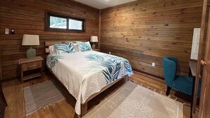 Un dormitorio con paredes de madera, un ventilador de techo, una cama con colcha estampada con hojas, dos mesitas de noche con lámparas, una ventana y una silla azul.